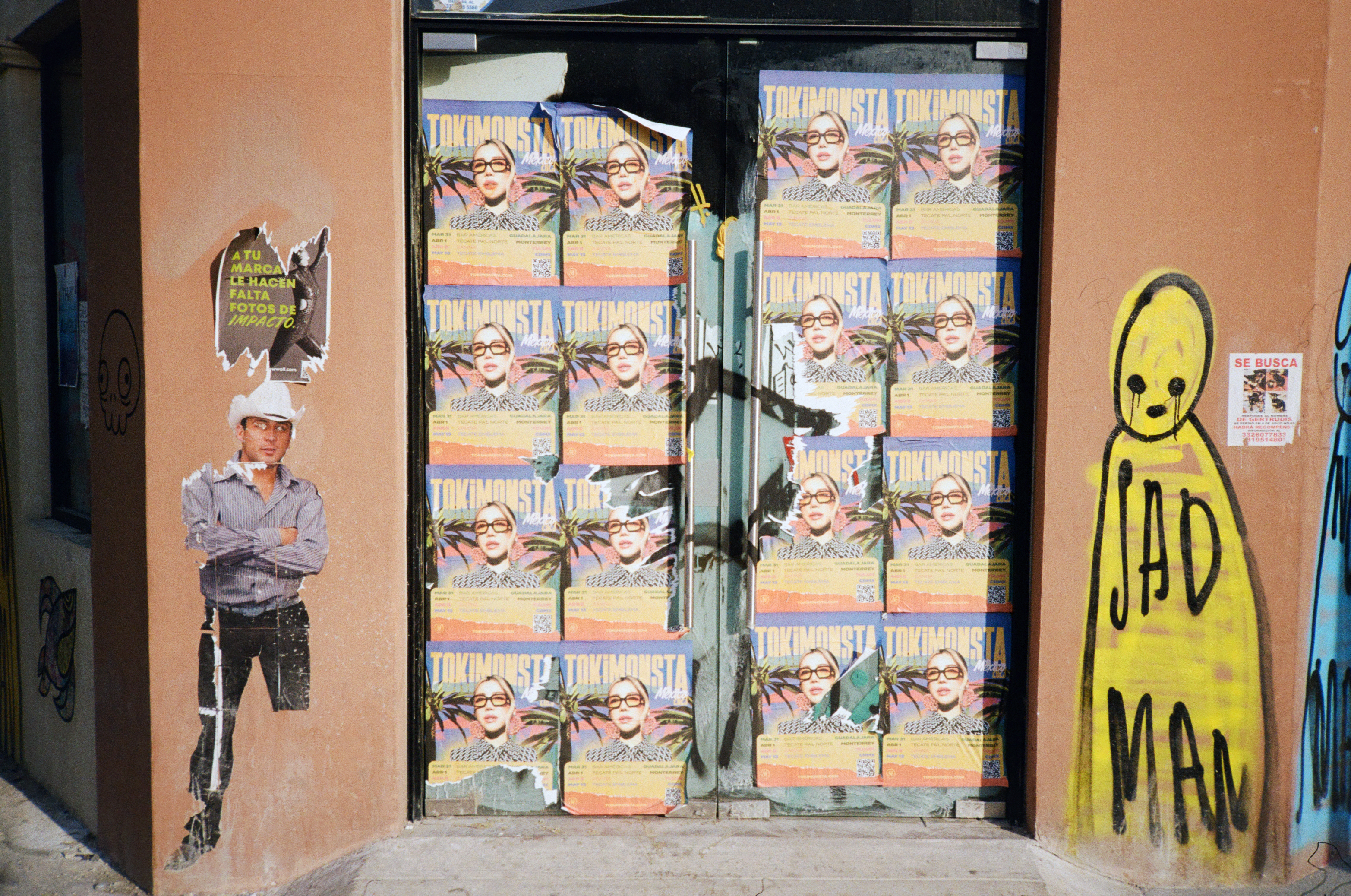 DOORS COVERED IN A GRID OF REPEATING WHEAT PASTED POSTERS WITH A WHEAT PASTED FIGURE OF A COWBOY ON THE LEFT AND A SPRAY PAINTED YELLOW FIGURE WITH THE TAG SAD MAN
