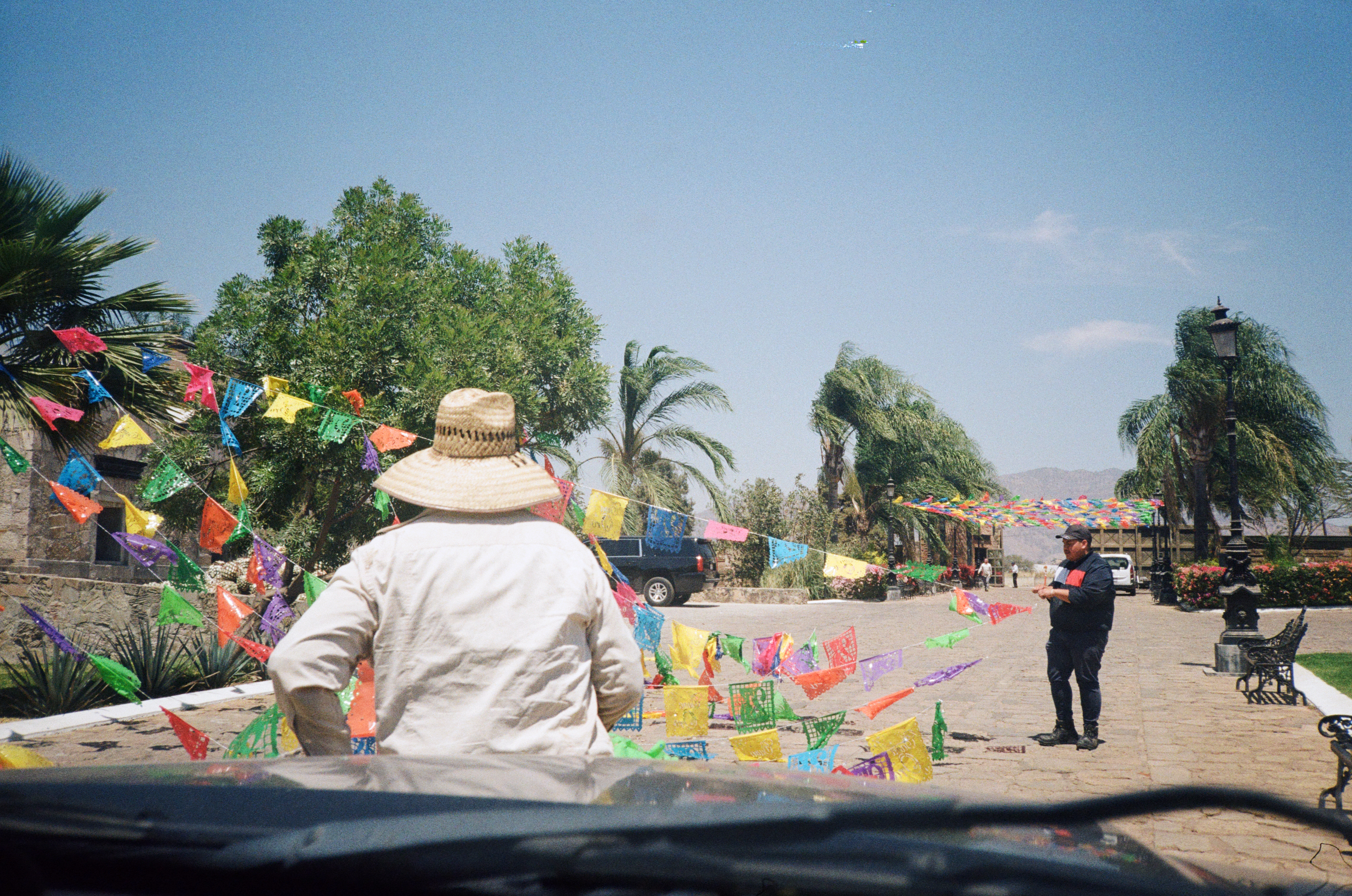 TWO MEN PUTTING UP BANNERS OF PAPEL PICADO PLASTICO IN THE DRIVEWAY OF A TEQUILA DISTILLERY, TAKEN FROM THE PASSENGER SEAT OF A CAR