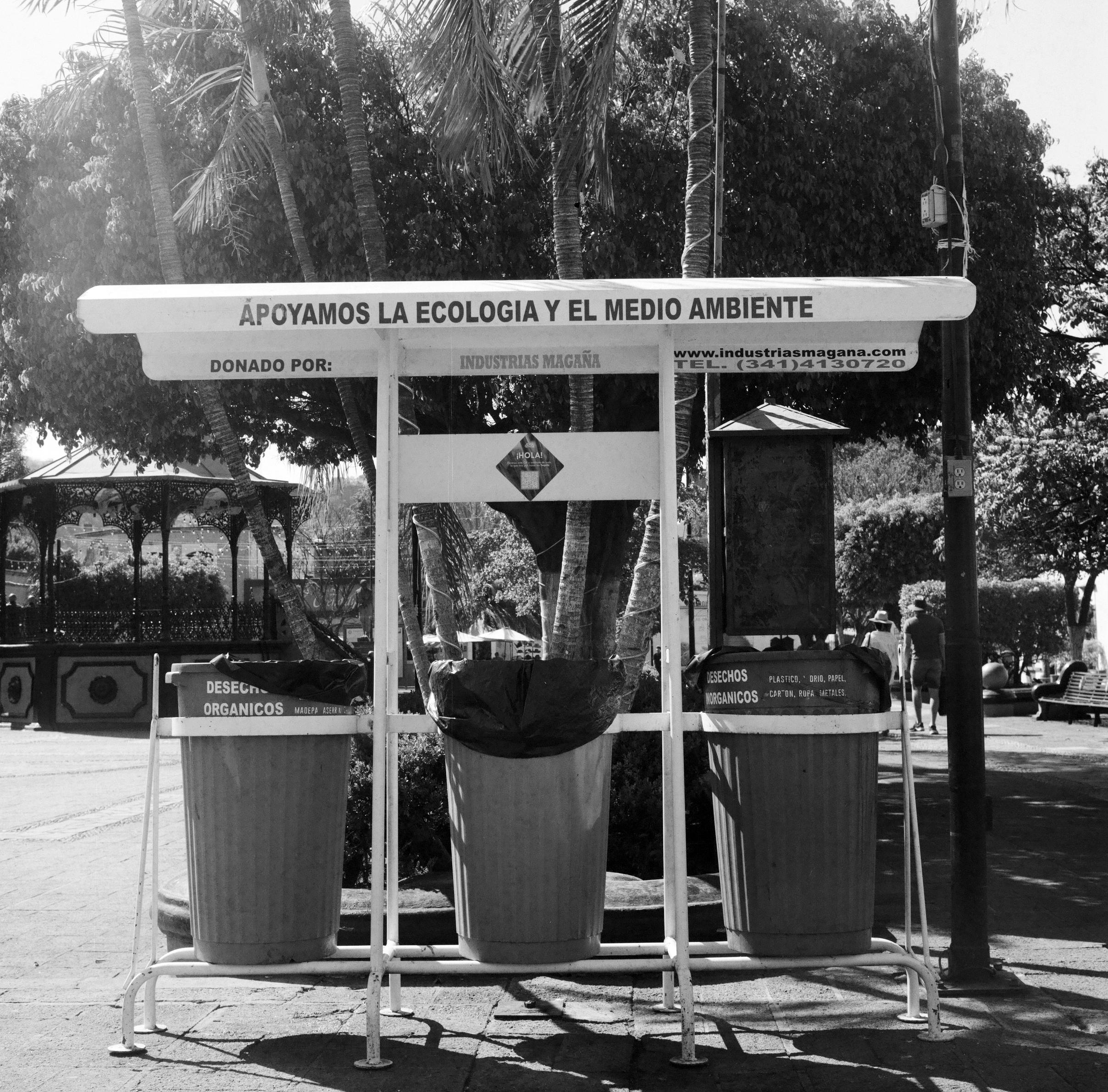 BLACK AND WHITE IMAGE OF GARBAGE & RECYCLING BINS WITH A SIGN THAT SAYS APOYAMOS LA ECOLOGIA Y EL MEDIO AMBIENTE