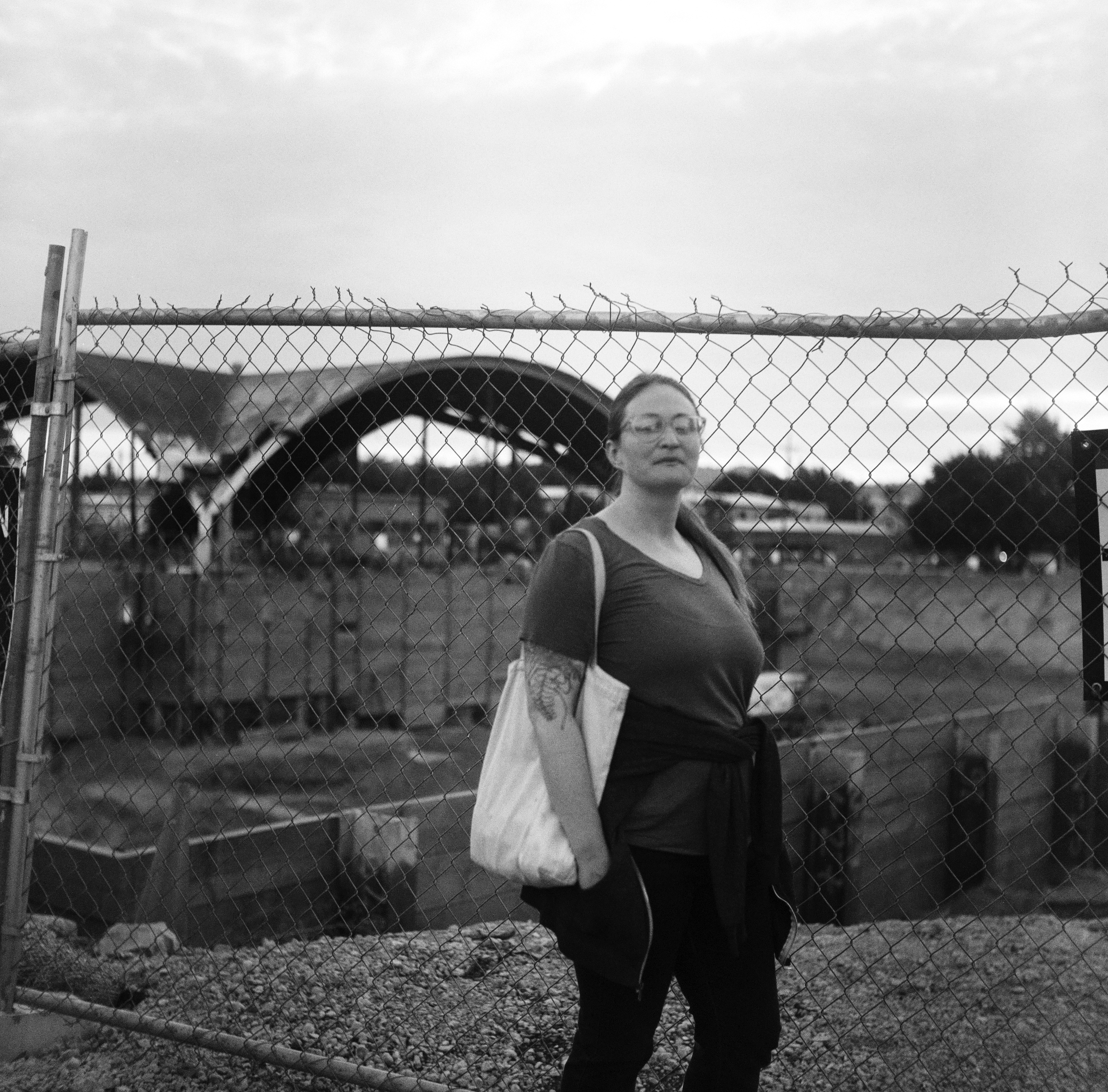 POPPY STANDS WITH A TOTE BAG OVER HER SHOULDER IN FRONT OF A CHAIN LINK FENCE AND THE OLD PEPSI ARCHWAY UNDER CONSTRUCTION ON SANDY BLVD