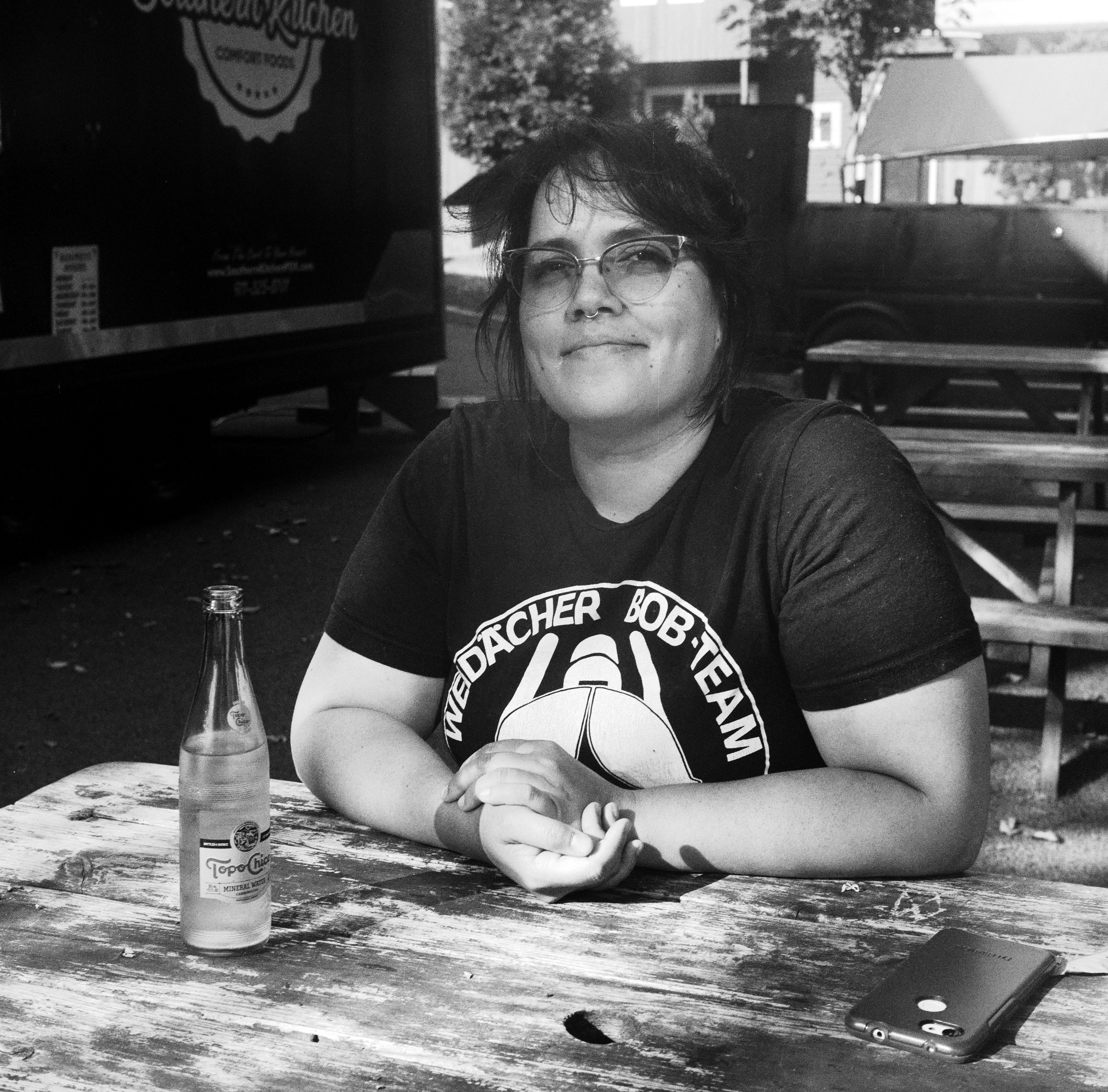 DANA SITS AT A PICNIC TABLE WITH A BOTTLE OF TOPO CHICO AND HER PHONE WHILE WAITING FOR SUNDAY MORNING FOOD AT KING BURRITO