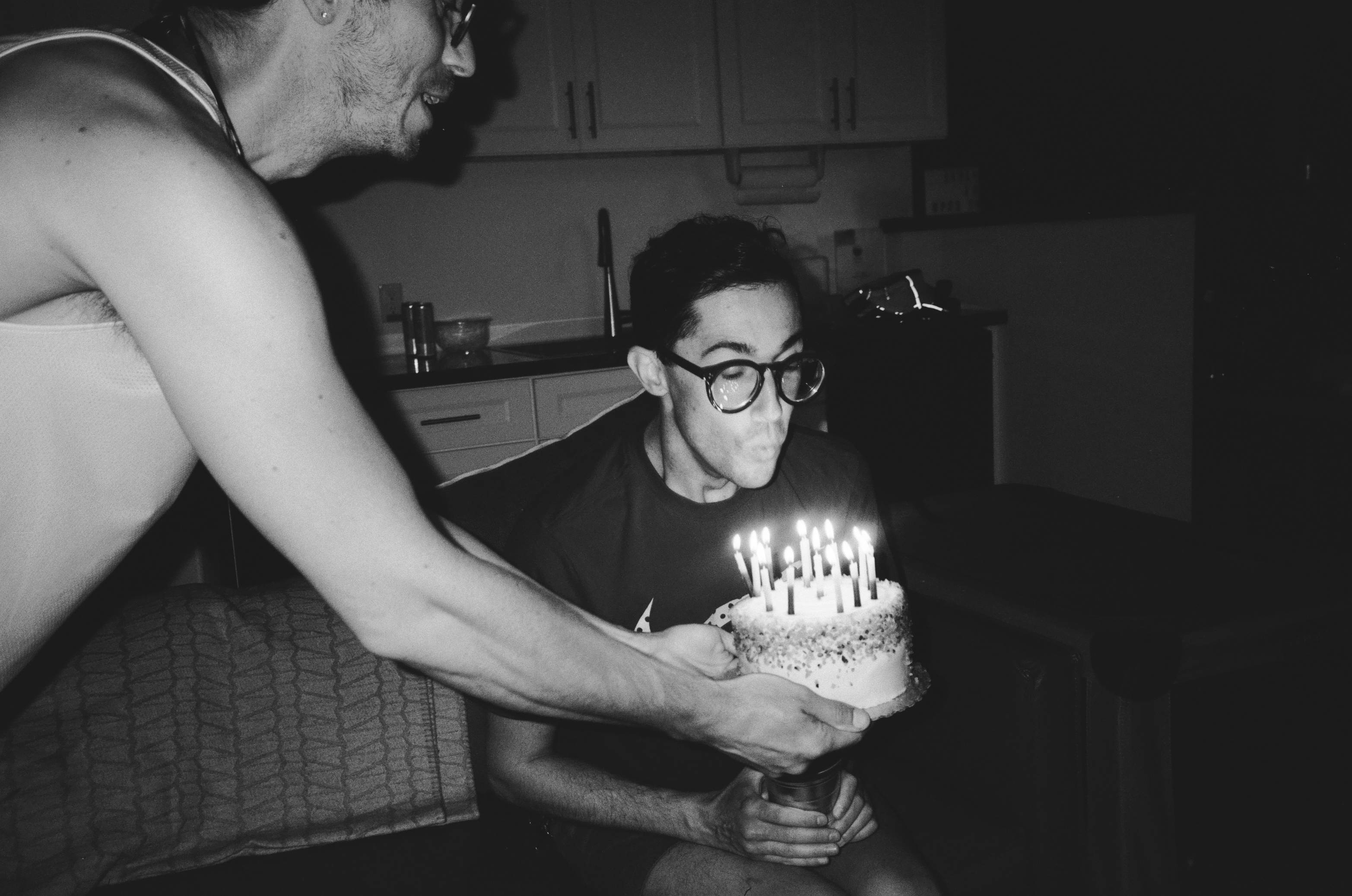 A MAN HOLDS A CAKE FULL OF CANDLES IN FRONT OF ANOTHER MAN WHO IS ABOUT TO BLOW THEM OUT