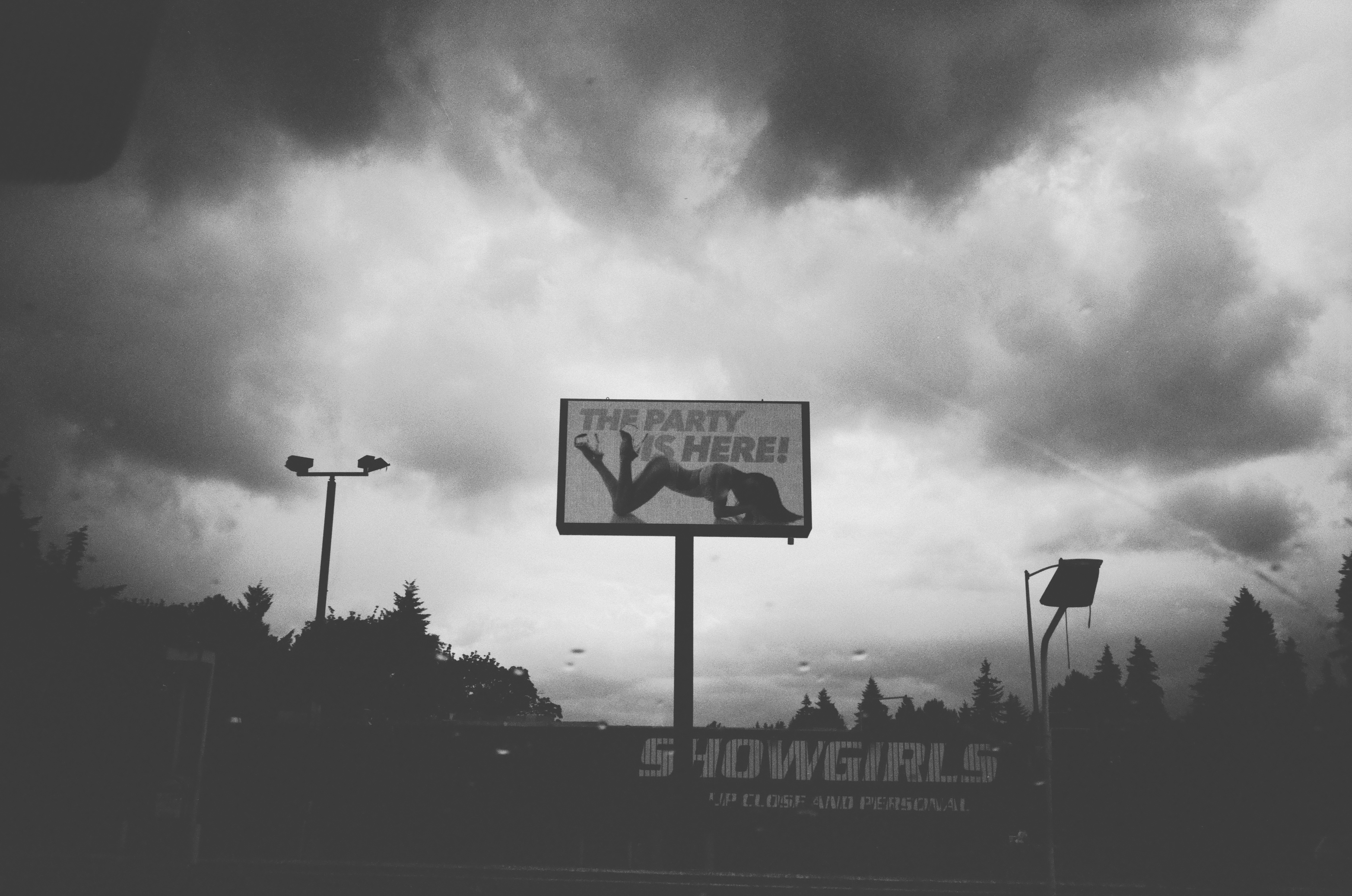 A BLACK AND WHITE IMAGE OF A LIGHT UP SIGN AT A STRIP CLUB THAT SAYS THE PARTY IS HERE AGAINST A GRAY AND RAINY SKY
