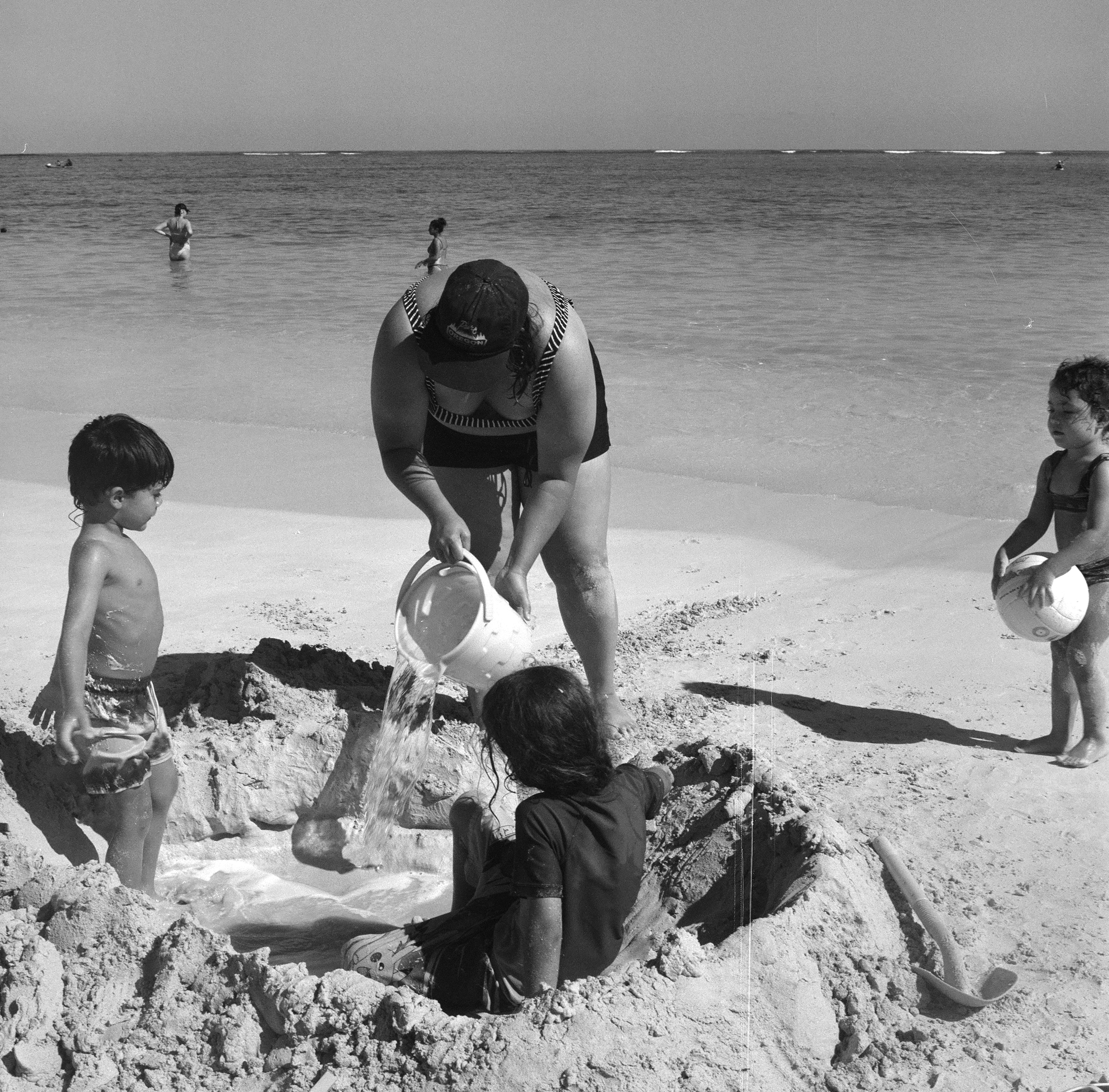 DANA POURS A BUCKET OF WATER INTO A MAJOR SAND CIRCLE THAT MICAH AND SONNY ARE INSIDE OF AS LYLA LOOKS ON HOLDING A VOLLEYBALL