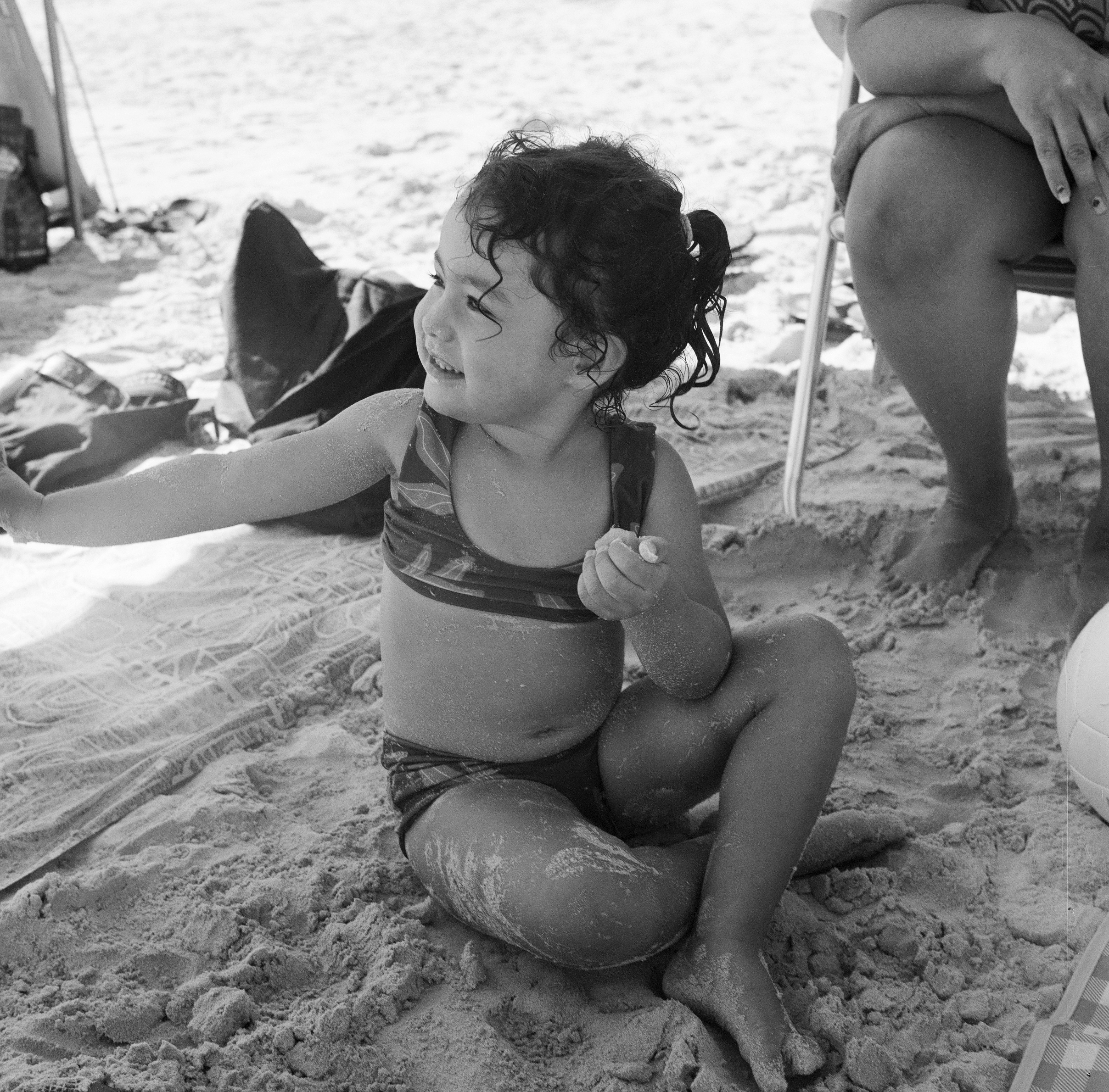 LYLA SITTING IN THE SAND UNDER THE SUN SHADE AT THE BEACH LOOKING OFF TO HER RIGHT AND SMILING