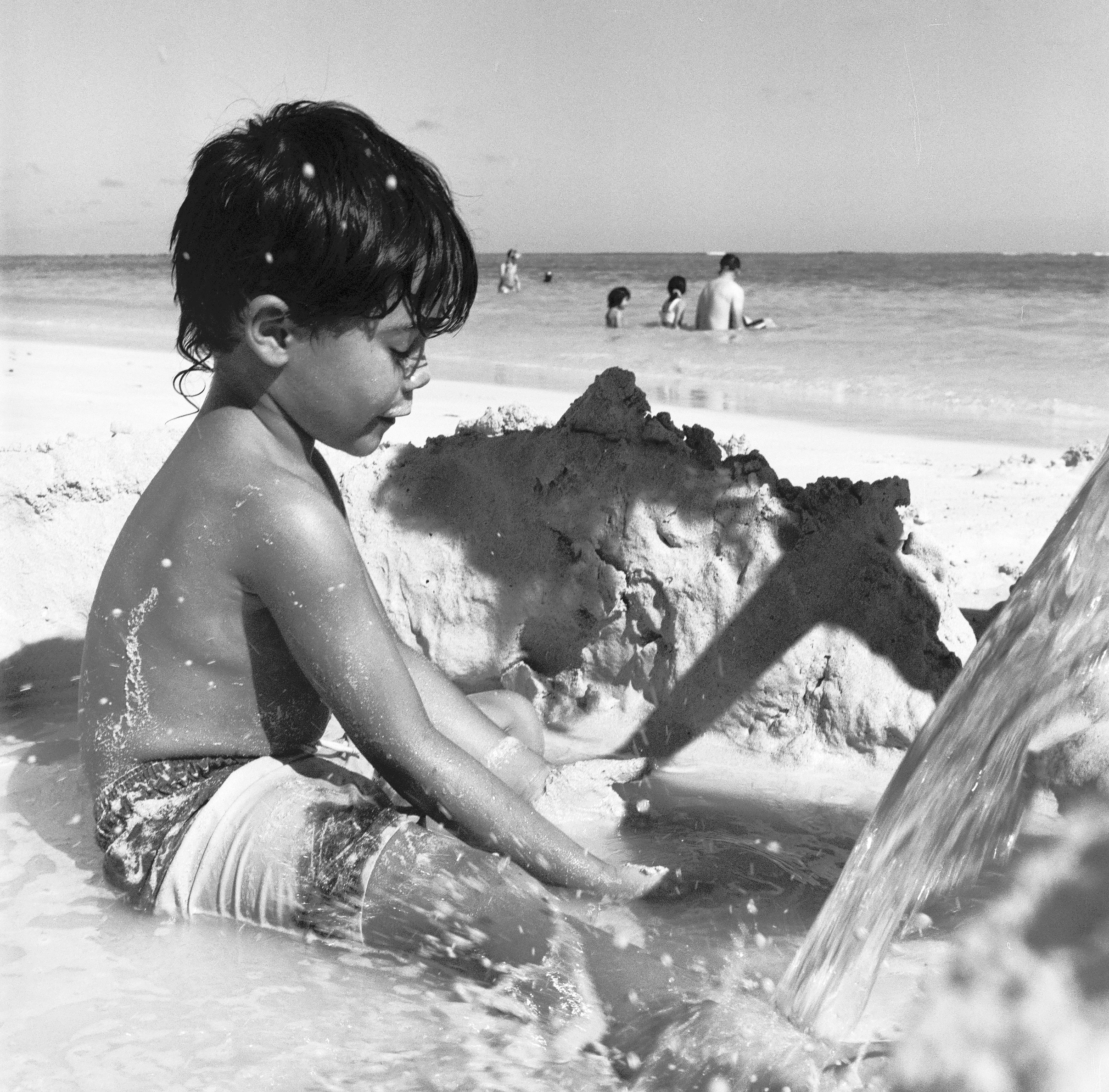 MICAH SITS IN A SAND BUILDING BEACH ZONE WHILE WATER POURS OUT OF A BUCKET AND SPLASHES SAND AROUND