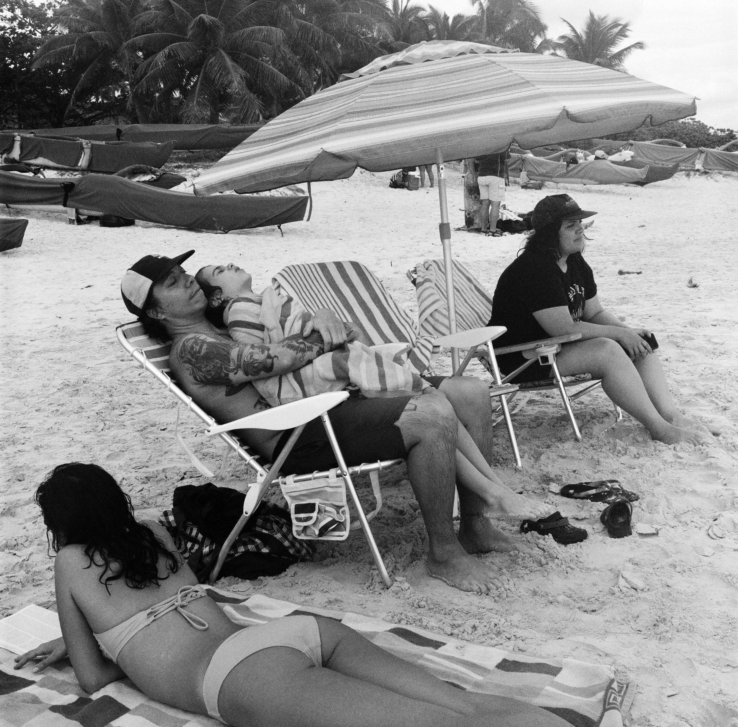 CHARLIE LAYS ON A BLANKET WHILE ALSTON HOLDS SONNY A TOWEL IN HIS LAP IN A FOLDING CHAIR AND DANA SITS IN ANOTHER CHAIR UNDER AN UMBRELLA