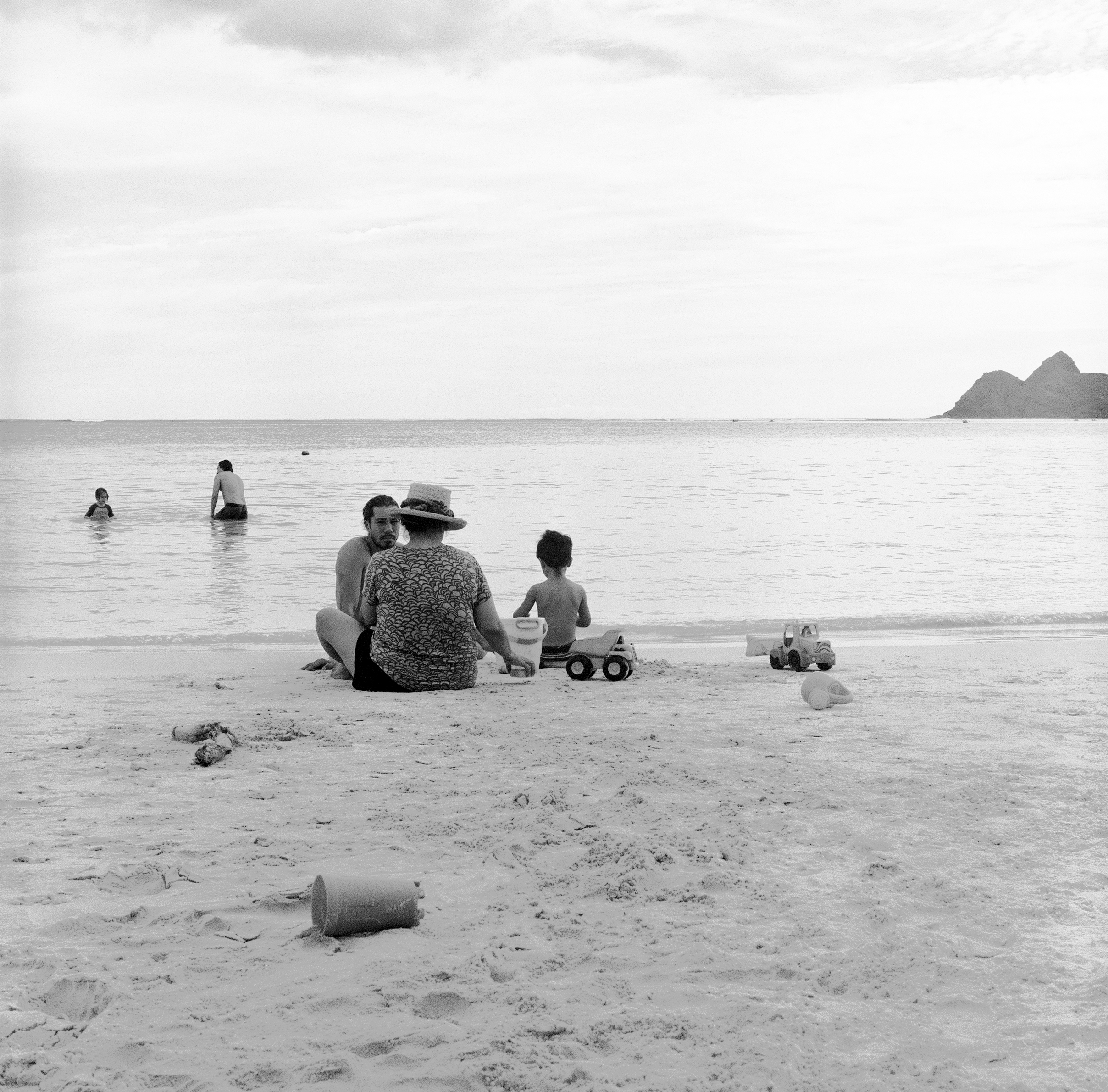 LINDSEY JAS AND MICAH SIT IN THE SAND NEAR THE WATER WHILE ALSTON AND SONNY PLAY IN THE WATER BEHIND