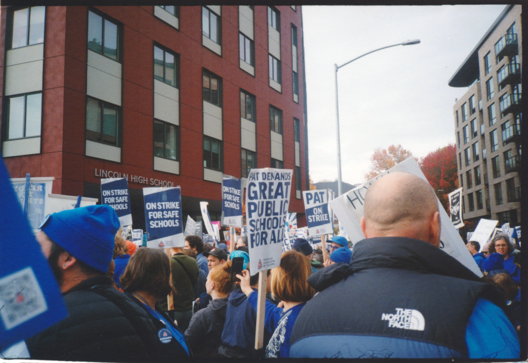 A COLOR PHOTO OF A CROWD OF PEOPLE IN BLUE HOLDING PICKET SIGNS IN FRONT OF A RED BUILDING THAT SAYS LINCOLN HIGH SCHOOL