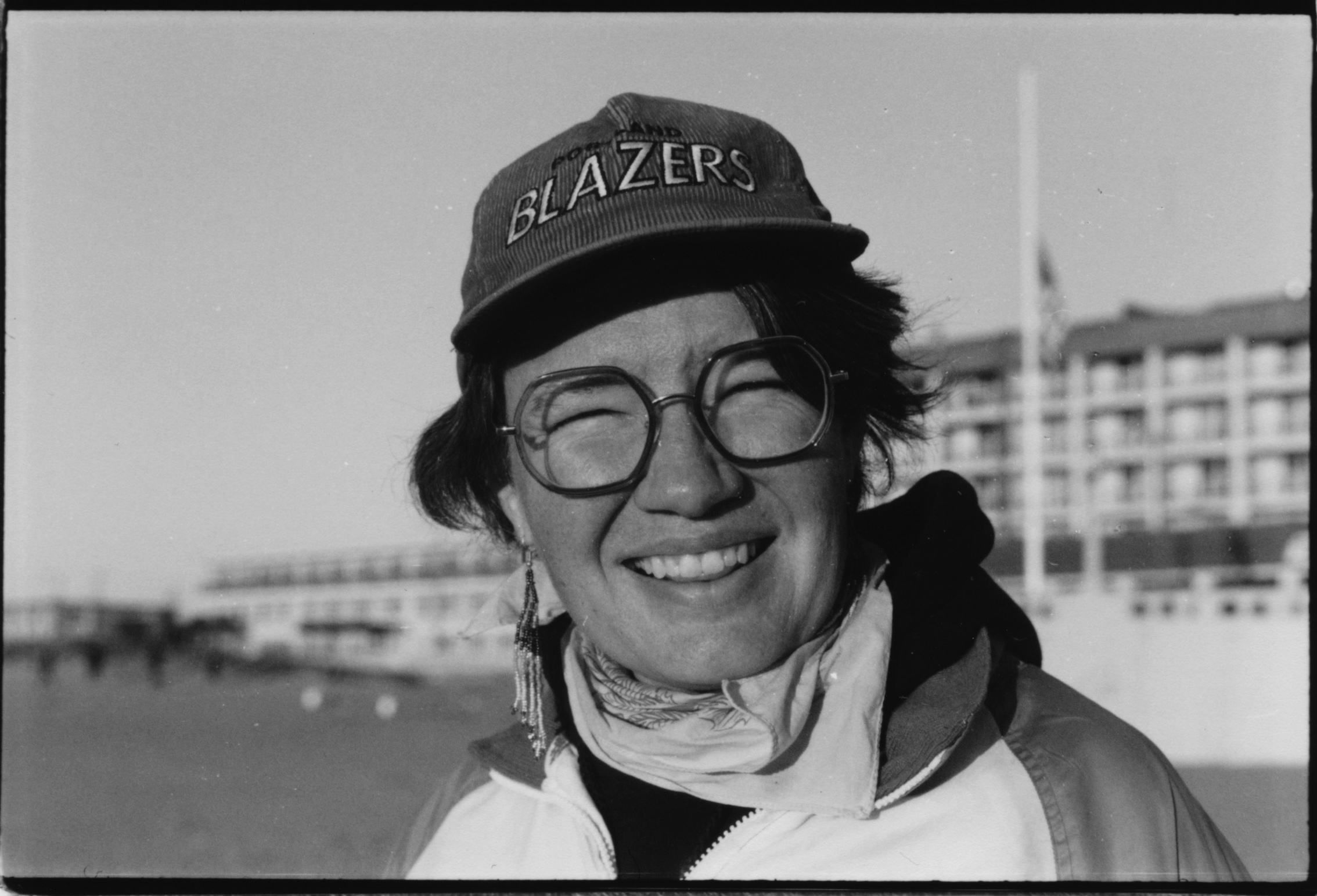 A BLACK AND WHITE PORTRAIT OF A WHITE LESBIAN IN A BLAZERS HAT AND GLASSES IN FRONT OF A BEACH HOTEL