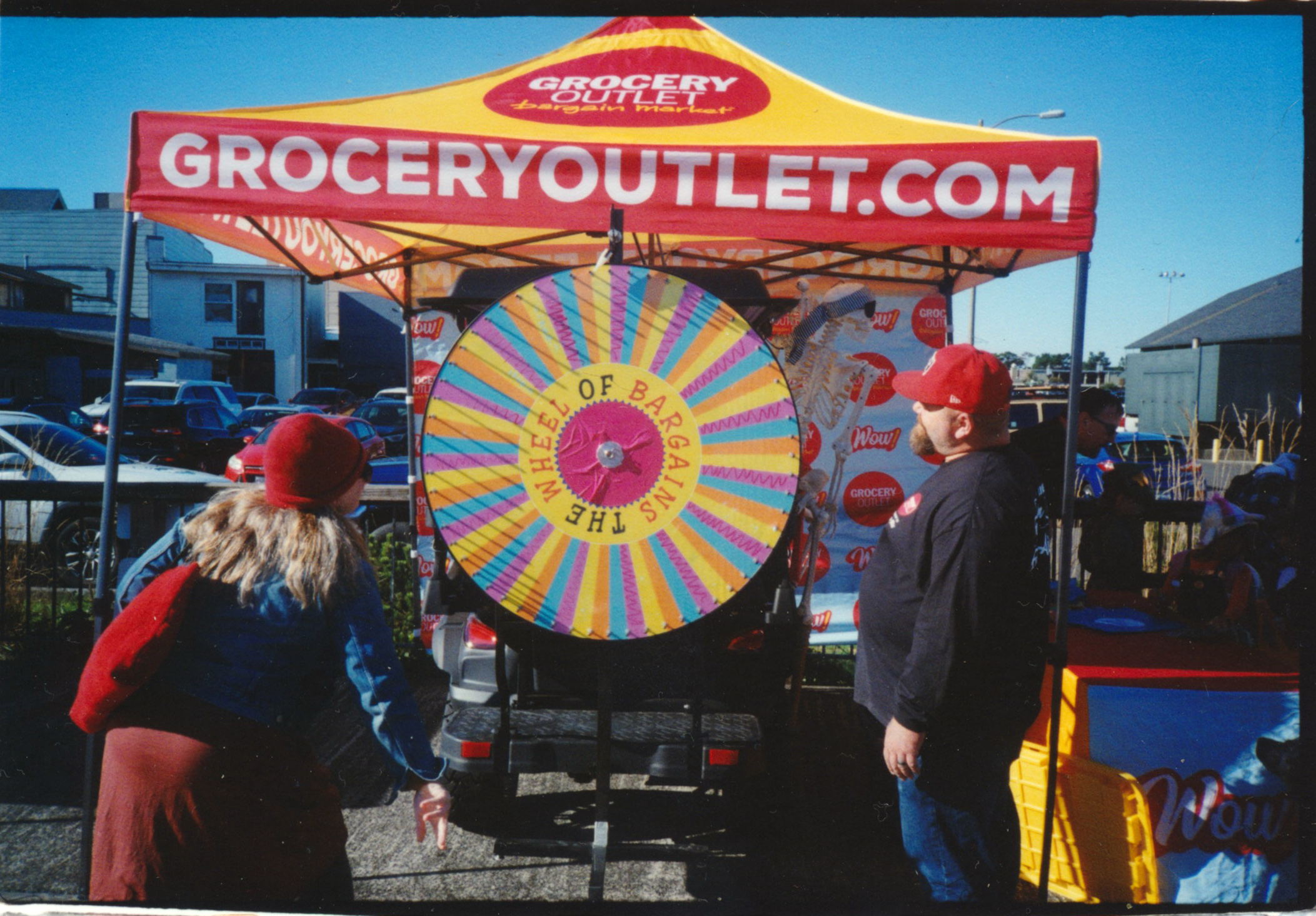 A WOMAN SPINS A WHEEL OF BARGAINS AT GROCERY OUTLET CANOPY AGAINST A BLUE SKY