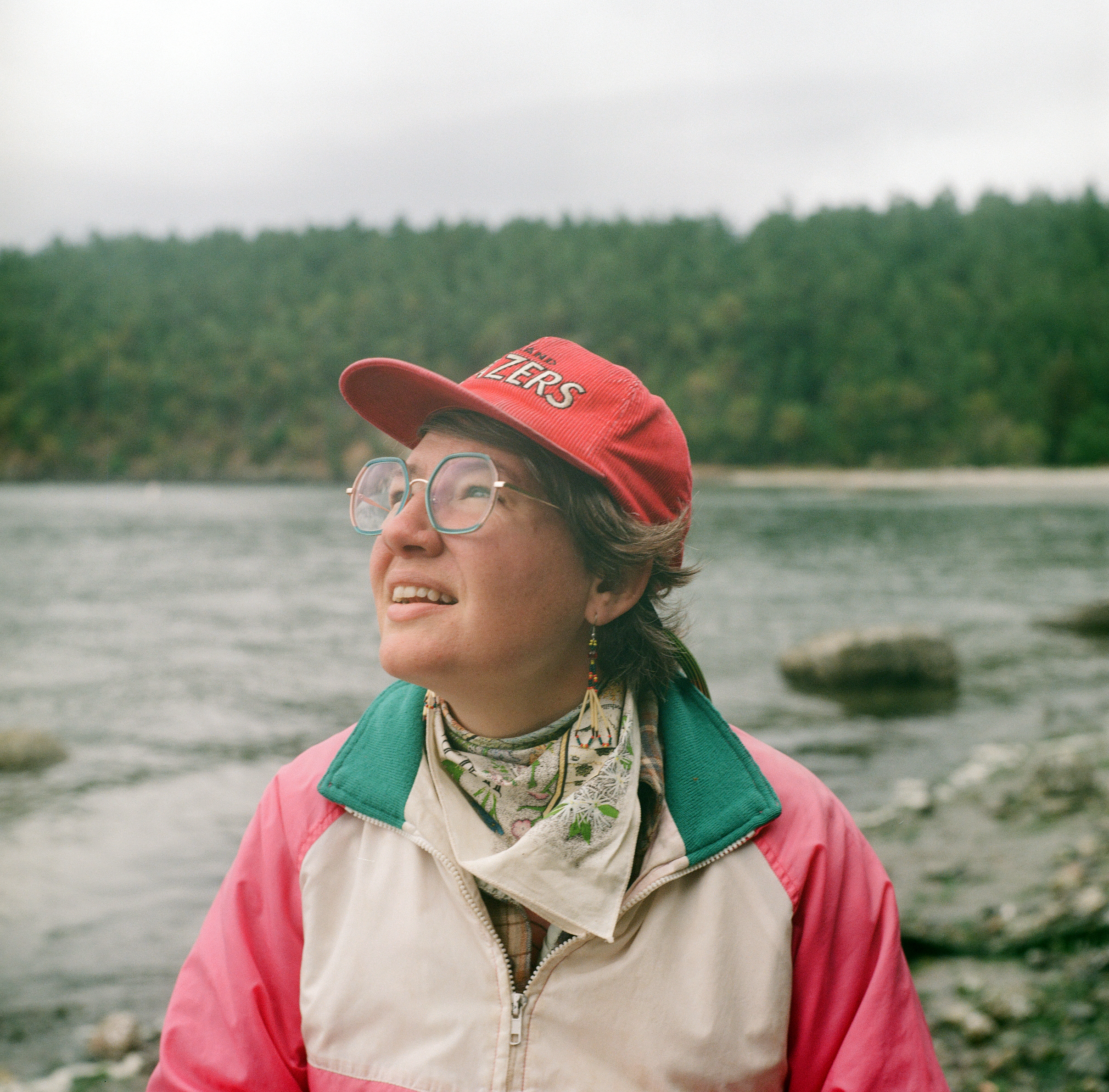 A WHITE LESBIAN IN A WINDBREAKER WITH A RED BLAZERS CORDUROY HAT AND A WILDFLOWERS BANDANA LOOKS UP INTO THE SKY WITH THE OCEAN COAST AND EVERGREEN TREES BEHIND HER