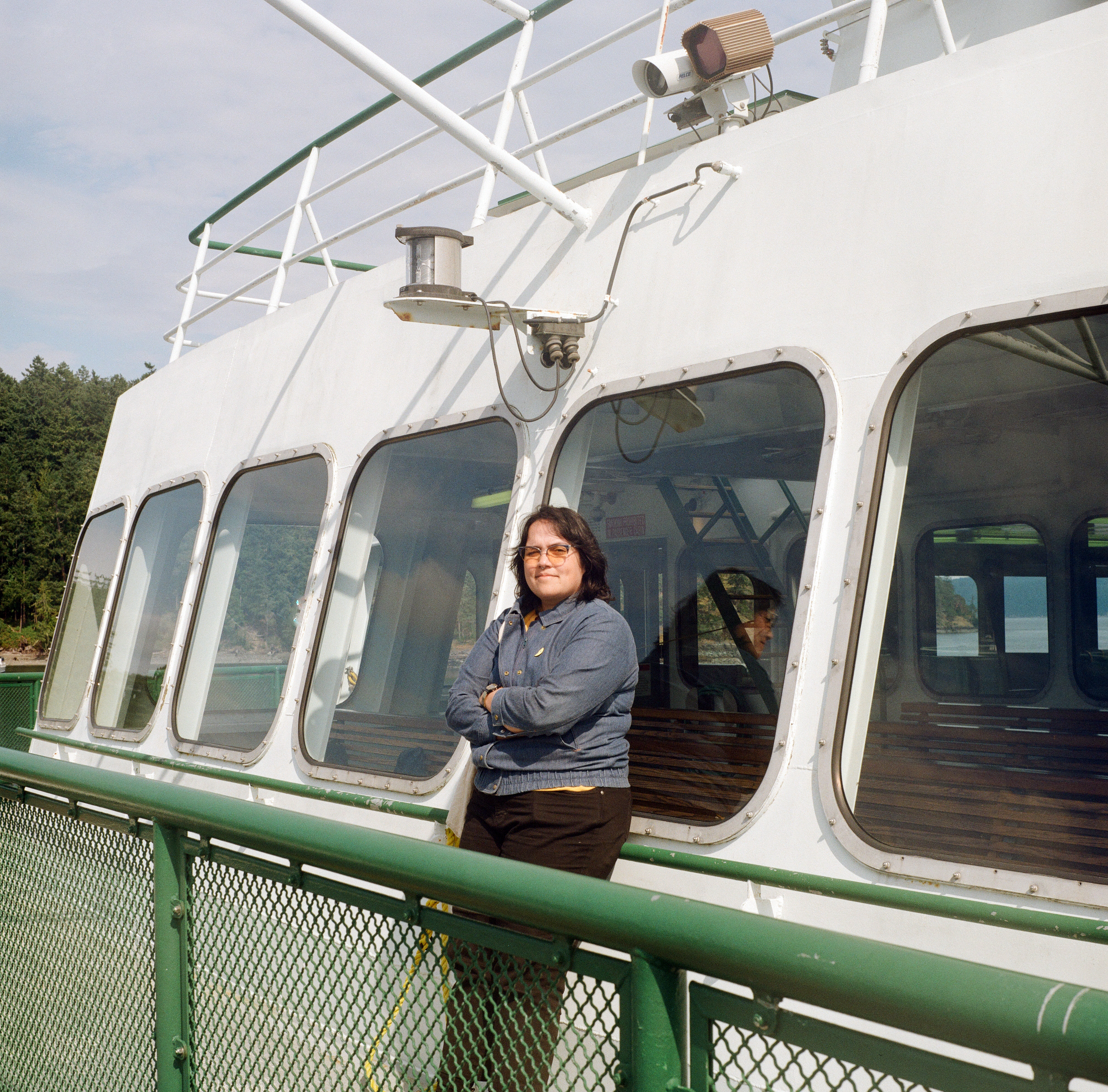 A HAWAIIAN PERSON IN A DENIM JACKET AND BLACK PANTS LEANS BACK AGAINST THE OBSERVATION WINDOW ON A WASHINGTON STATE FERRY AND CROSSES THEIR ARMS