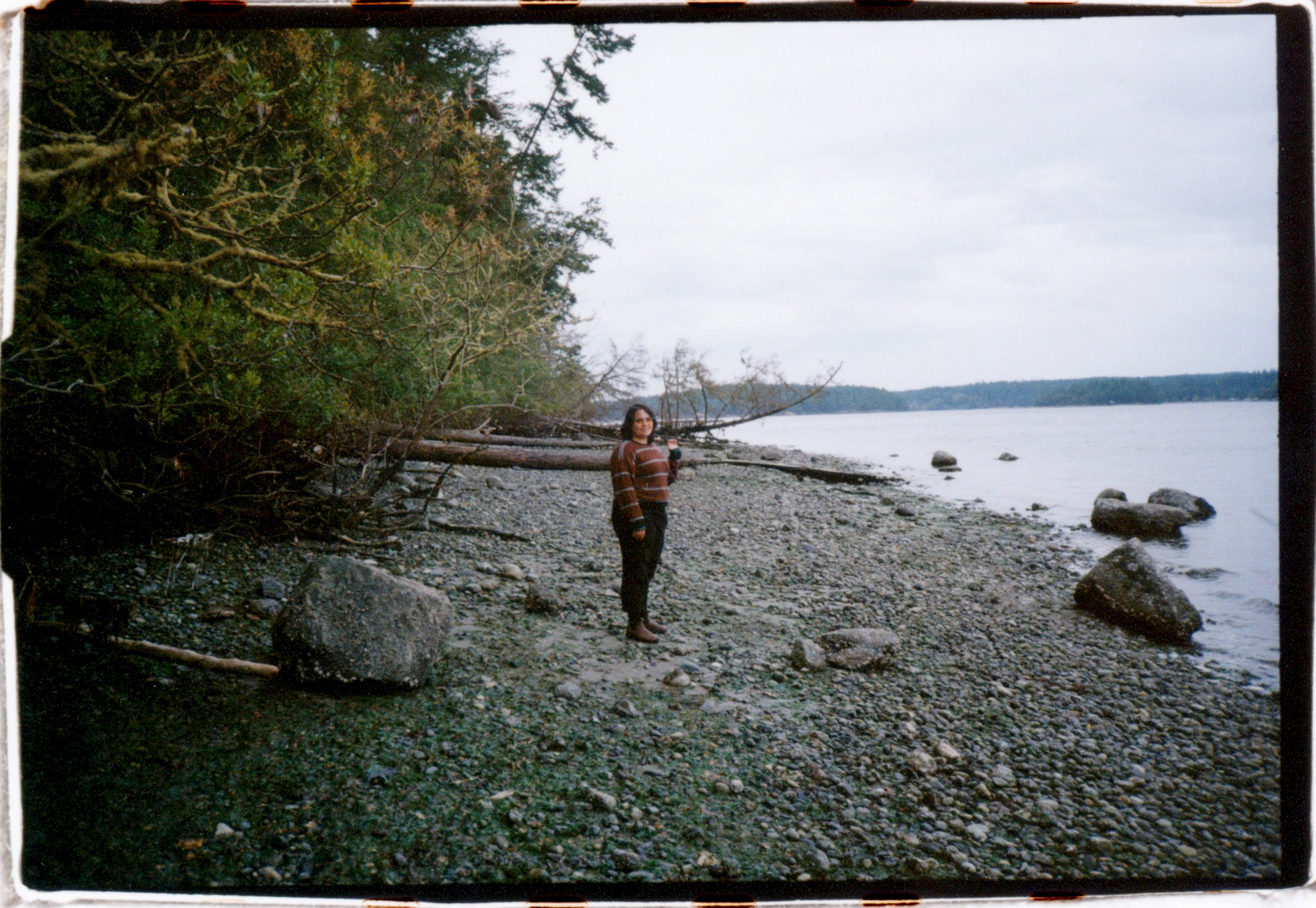 A PERSON ON A ROCKY BEACH TURNING TO FACE THE CAMERA WITH WILLOWS AND TREES BEHIND THEM
