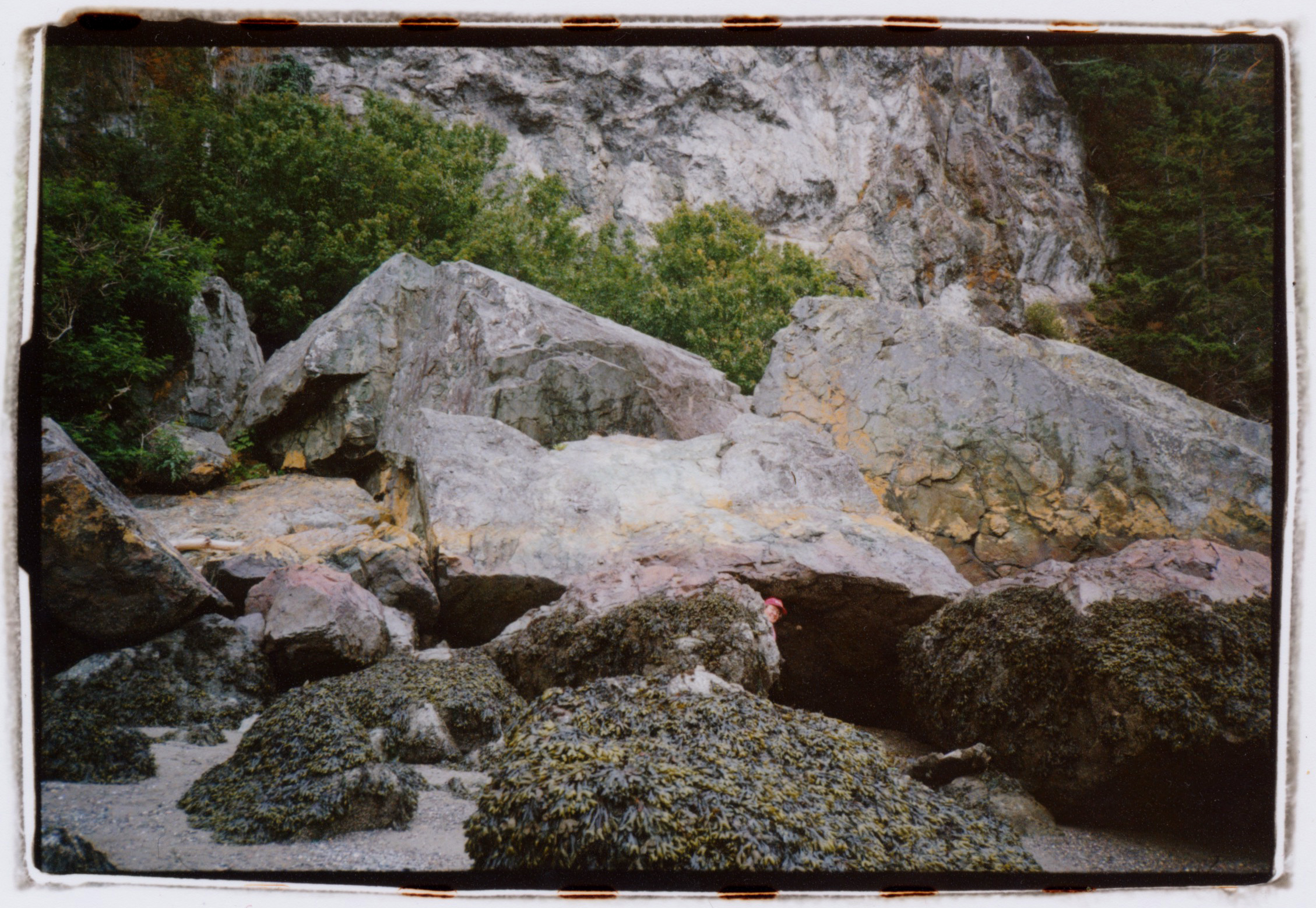 AN IMAGE OF ENORMOUS BOULDERS AT THE BASE OF A CLIFF NEXT TO THE OCEAN WITH A PERSON PEEPING OUT FROM BEHIND ONE