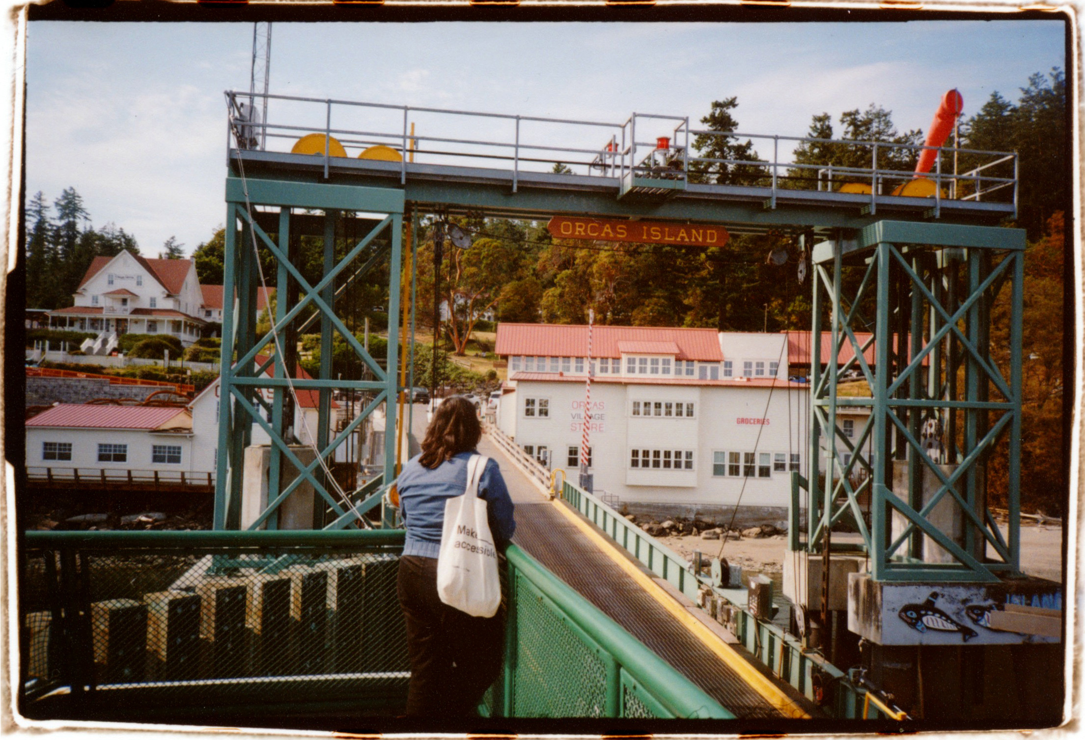 A PERSON HOLDING A WHITE TOTE BAG STANDS ON THE DECK OF A FERRY LOOKING AT THE ORCAS ISLAND TERMINAL